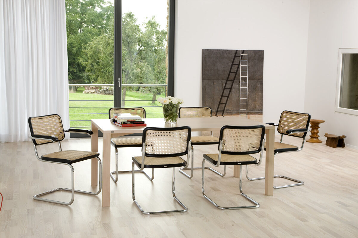 Modernes Esszimmer mit hellbraunem Holztisch und sechs Thonet S 32 Stahlrohr-Freischwinger Stühlen in Schwarz und Beige. Große Fenster mit Blick auf grüne Landschaft im Hintergrund. Stilvolles und minimalistisches Design.