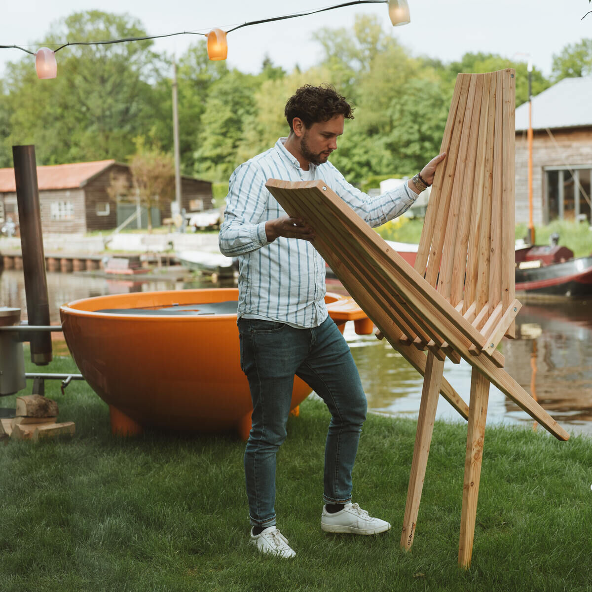 Mann stellt eine Holzliege im Garten auf, umgeben von grünem Rasen, einem Wasserkanal im Hintergrund und beleuchteten Lampen. Eine orangefarbene Badewanne und ein kleiner weißer Tisch mit Gläsern sind ebenfalls sichtbar.