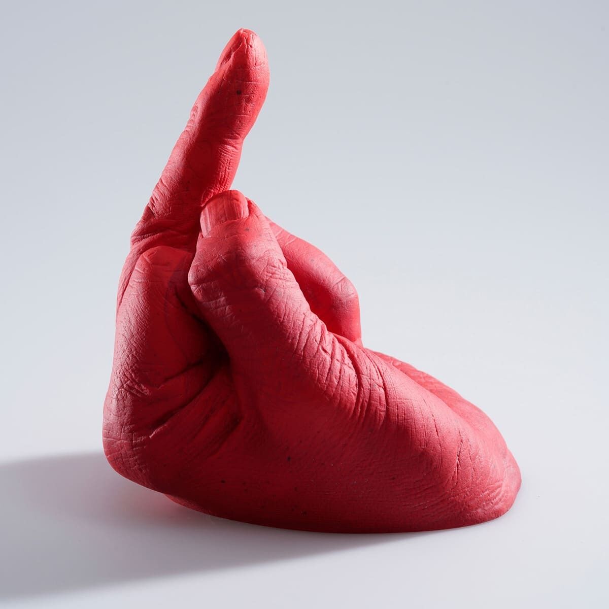 Eine rote Skulptur eines nach oben gestreckten Mittelfingers, ein Kunstwerk von Ai Weiwei aus der Serie »Study of Perspective in Glass«. Die Skulptur hat eine detaillierte Textur und steht auf einer weißen Oberfläche, wobei sie Schatten wirft.