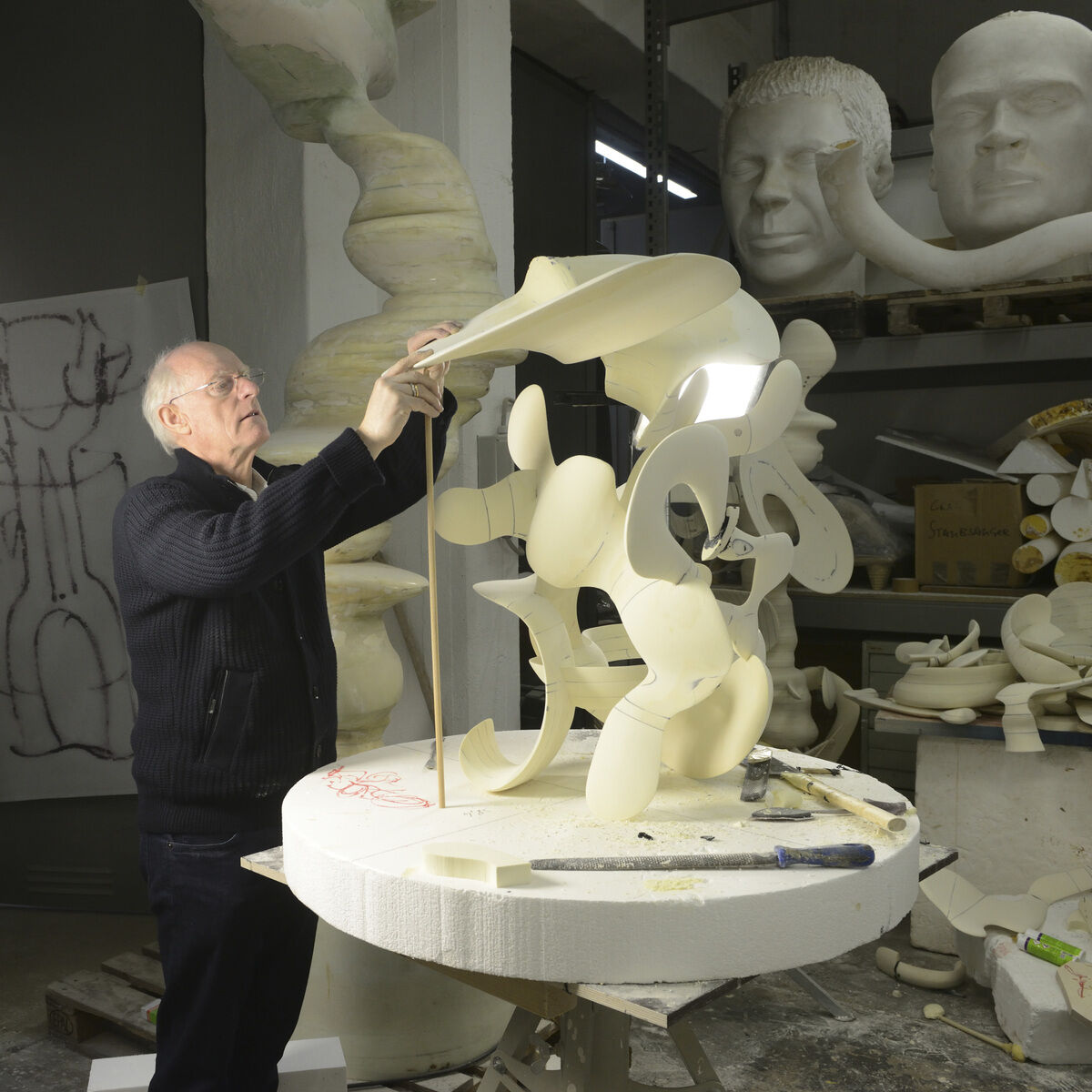 Ein älterer Mann arbeitet an einer abstrakten Skulptur in einem Studio, das mit verschiedenen Kunstwerken und großen Gesichtsskulpturen dekoriert ist.