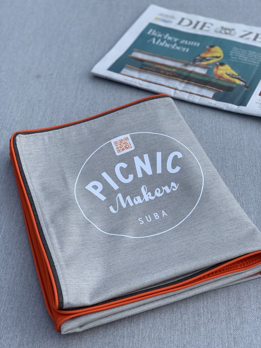 Eine graue Picknickdecke mit orangenen Rändern und der Aufschrift 'PICNIC Makers SUBA'. Im Hintergrund liegt eine Zeitung, teilweise sichtbar. Die Decke ist auf einer grauen Fläche ausgebreitet.