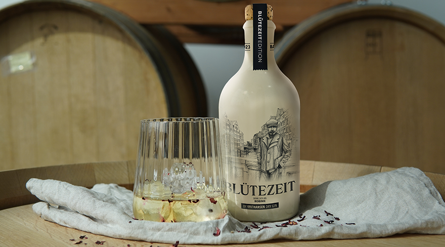 Weiße Flasche Blütezeit Dry Gin mit gezeichneter Mann-Skizze neben Glas mit Eiswürfeln auf Holzfass und grauem Tuch