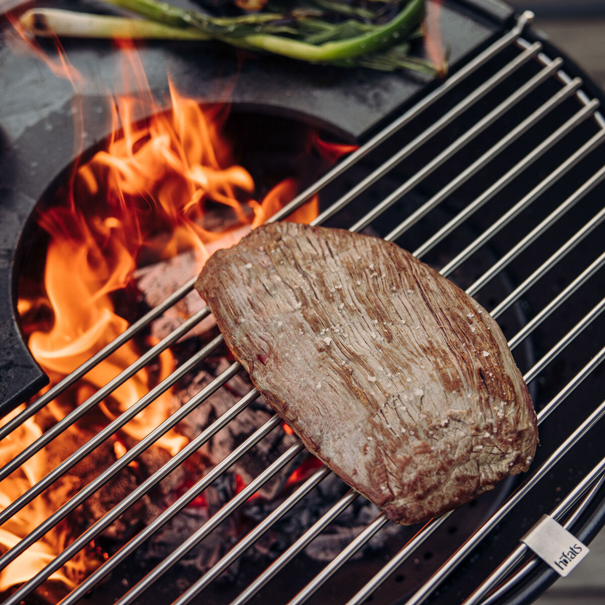 Ein Edelstahl-Grillrost des Produkts '»BOWL« Grillrost Edelstahl' mit darauf gegrilltem Fleisch und Gemüse. Im Hintergrund lodert ein Feuer in der Grillkugel. Die Aufnahme zeigt eine Outdoor-Kochsituation mit deutlichen Flammen und Holzkohle.