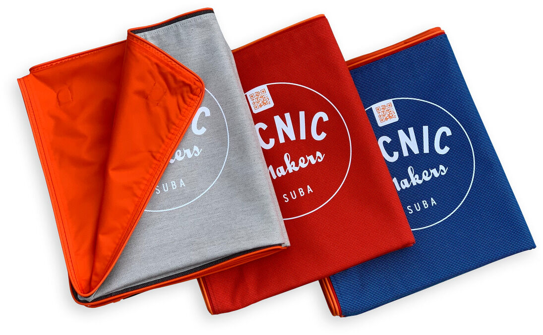 Drei Picknickdecken mit der Aufschrift ‚PICNIC makers‘ in Orange, Grau, Rot und Blau. Die Decken sind zusammenklappbar und aus robustem, wasserdichtem Material.
