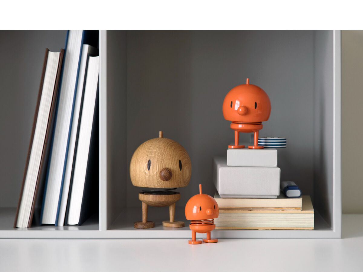 Drei Hoptimist Bumble-Figuren aus Eiche und orangefarbenem Kunststoff stehen in einem minimalistischen Regal, umgeben von Büchern und anderen kleinen Dekorationsobjekten.