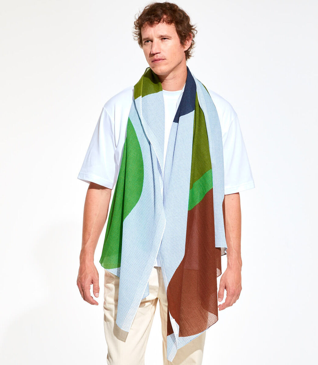 Ein Mann trägt ein Baumwolltuch namens 'Galet', das in Blau-, Grün- und Brauntönen gemustert ist. Das Tuch hängt lose um seine Schultern und wird über einem weißen T-Shirt und einer hellen Hose getragen.