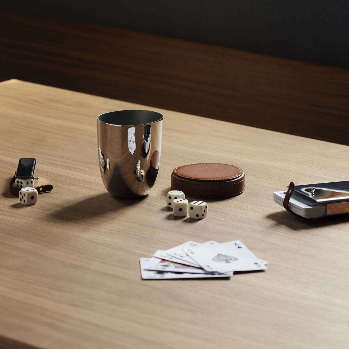 Ein metallischer Würfelbecher, ein Kartenstapel, einige Würfel und ein braunes Lederetui liegen auf einem Holztisch.