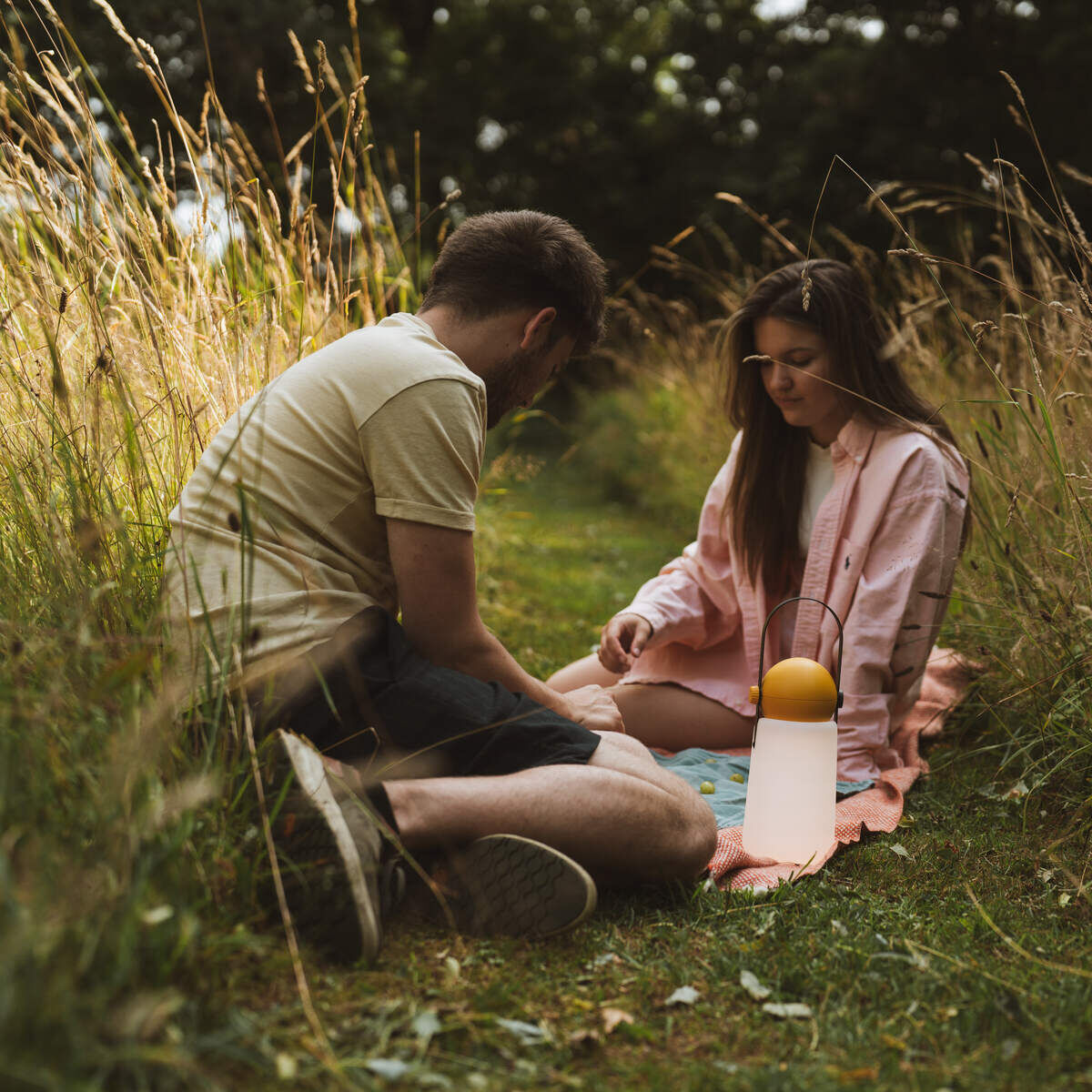 Eine portable Lampe namens »Guidelight« steht auf einer Wiese neben sitzenden Personen im Freien. Die Szene vermittelt eine gemütliche und entspannte Atmosphäre, ideal für Outdoor-Abende.