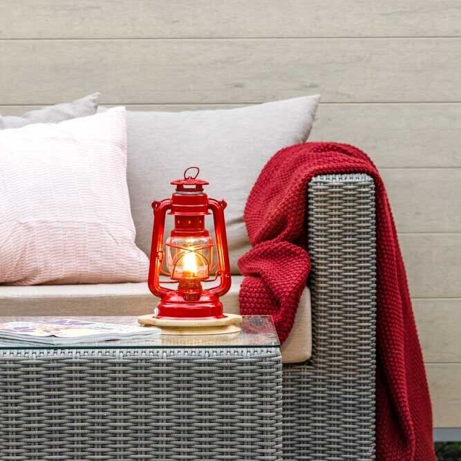Ein Wohnbereich mit grauen Rattan-Möbeln, dekoriert mit rot-weißen Kissen und einer roten LED-Laterne auf einem Tisch, die eine warme, gemütliche Beleuchtung ausstrahlt.