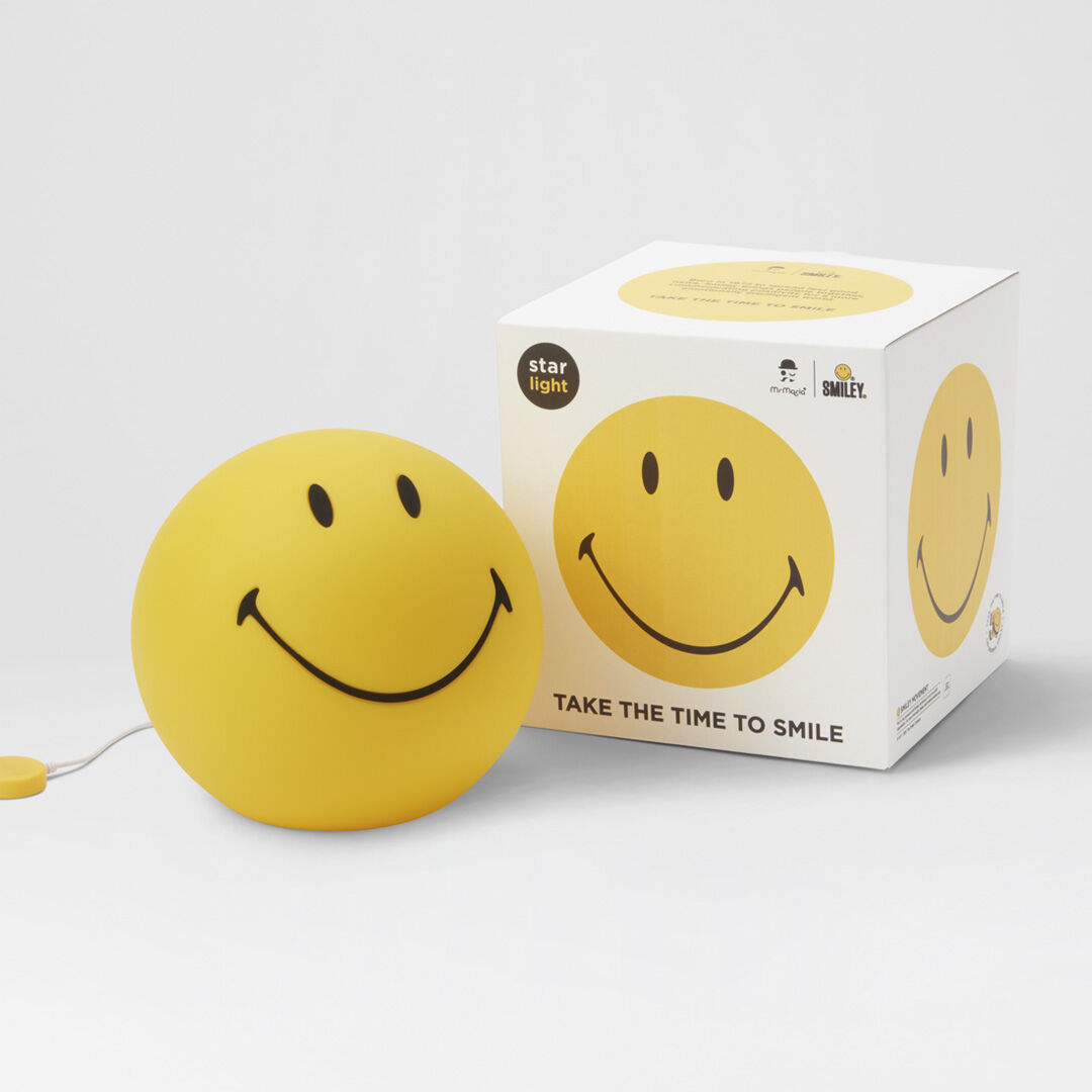 Abgebildet ist eine runde, gelbe LED-Lampe mit einem aufgemalten Smiley-Gesicht, daneben ist die passende Verpackung zu sehen. Die Lampe steht auf einer weißen Oberfläche.