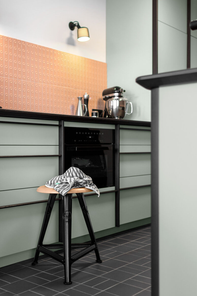 Modernes Kücheninterieur mit einem Rowac-Schemel in Eiche Mattschwarz im Vordergrund, umgeben von mintgrünen Schränken, einer Arbeitsfläche und einer mintgrünen Wand mit terrakottafarbenen Fliesen.