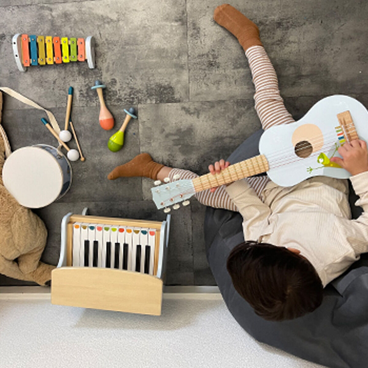 Ein Kind sitzt auf einem grauen Sitzsack und spielt in einem Zimmer mit einem Musik-Set bestehend aus einer kleinen Gitarre, einem Keyboard, einem Xylophon, einer Trommel mit Drumsticks und Maracas in verschiedenen Farben. Neben dem Kind liegt ein großes, hellbraunes Kuscheltier auf einem dunklen Boden.
