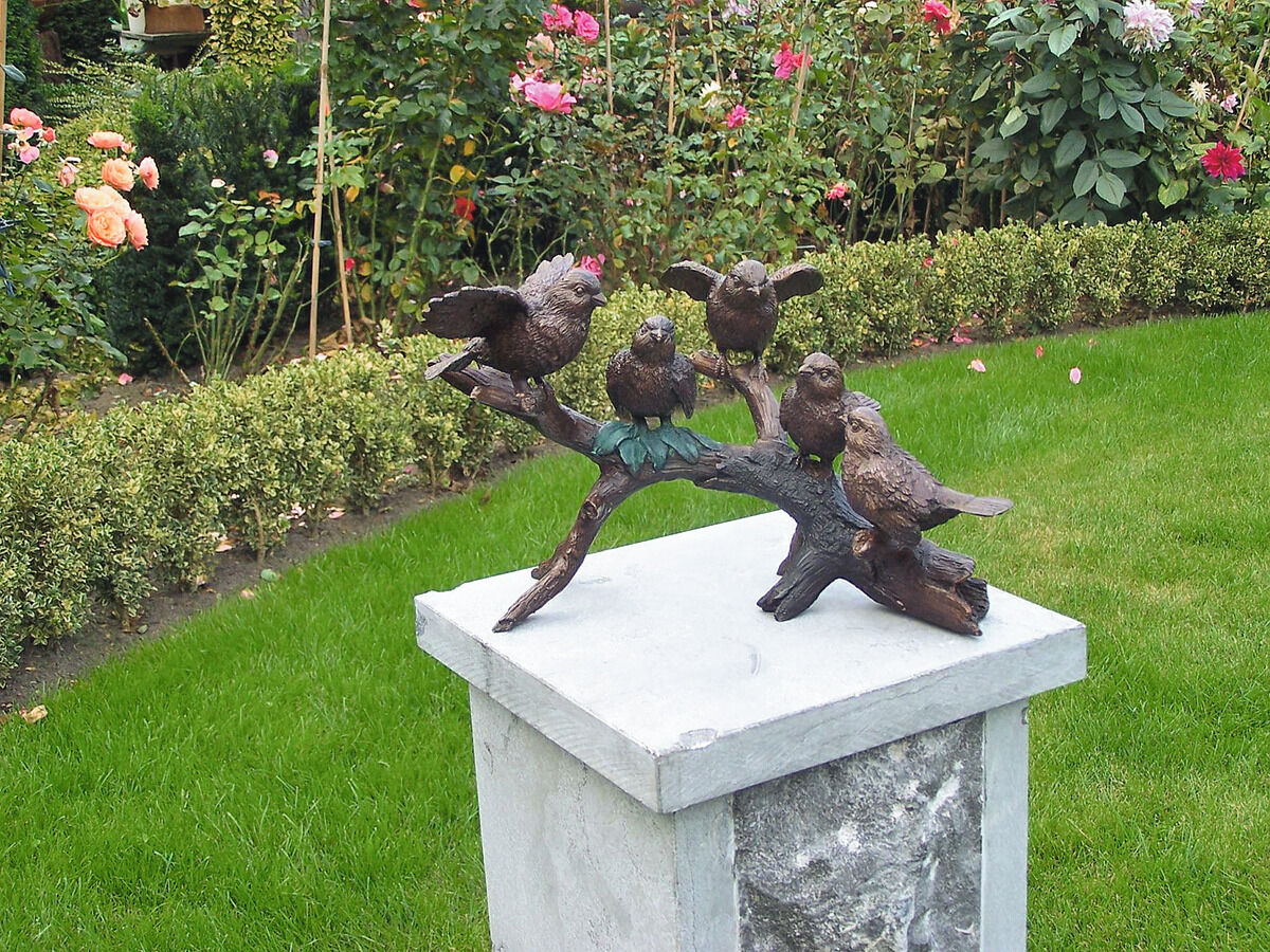 Gartenskulptur »Vögel auf Ast«, zeigt fünf kleine Vögel, die auf einem Ast sitzen. Die Skulptur steht auf einem steinernen Sockel in einem blühenden Garten mit grünen Hecken und bunten Blumen.