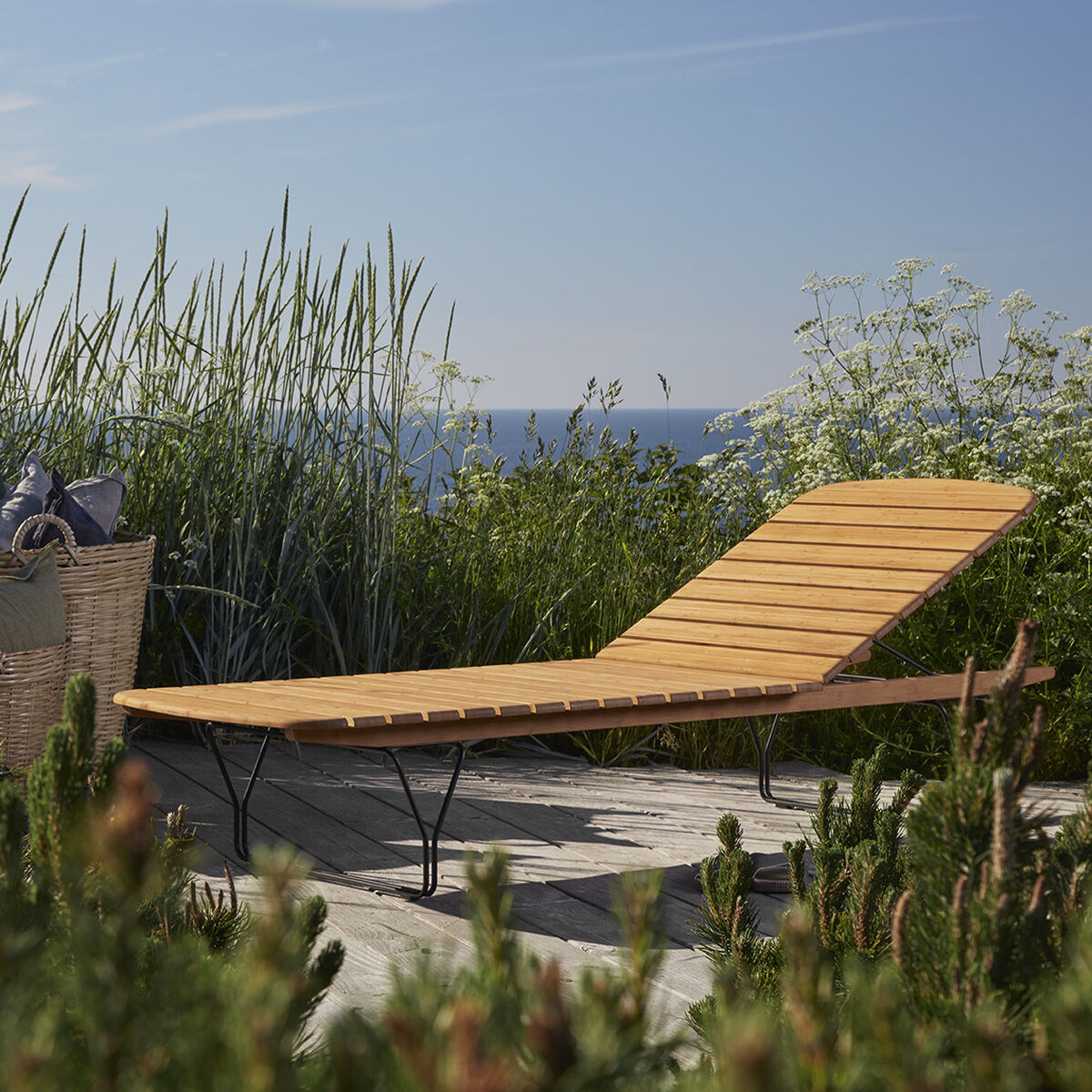 Eine moderne Holzliege von MOLO steht auf einer Terrasse im Freien mit Blick auf das Meer. Im Hintergrund sind hohe Gräser und ein klarer blauer Himmel zu sehen. Neben der Liege stehen Körbe mit Textilien.