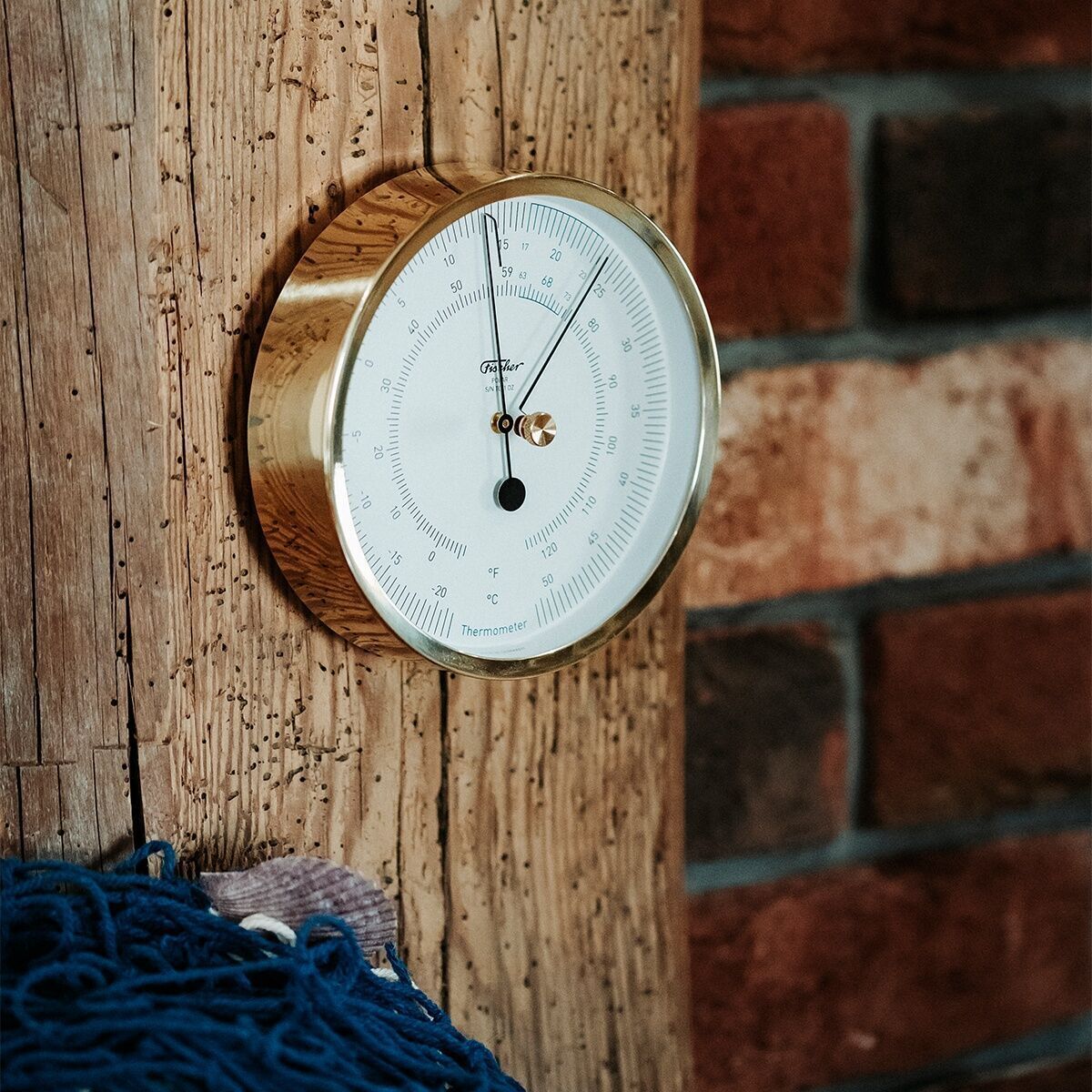Ein ZEIT-Sonderedition »POLAR Thermometer« an einer rustikalen Holzwand montiert. Der runde Rahmen des Thermometers ist golden, das Zifferblatt ist weiß mit feinen Markierungen und zwei schwarzen Zeigern. Im Hintergrund sind Holz- und Ziegelwände zu sehen.