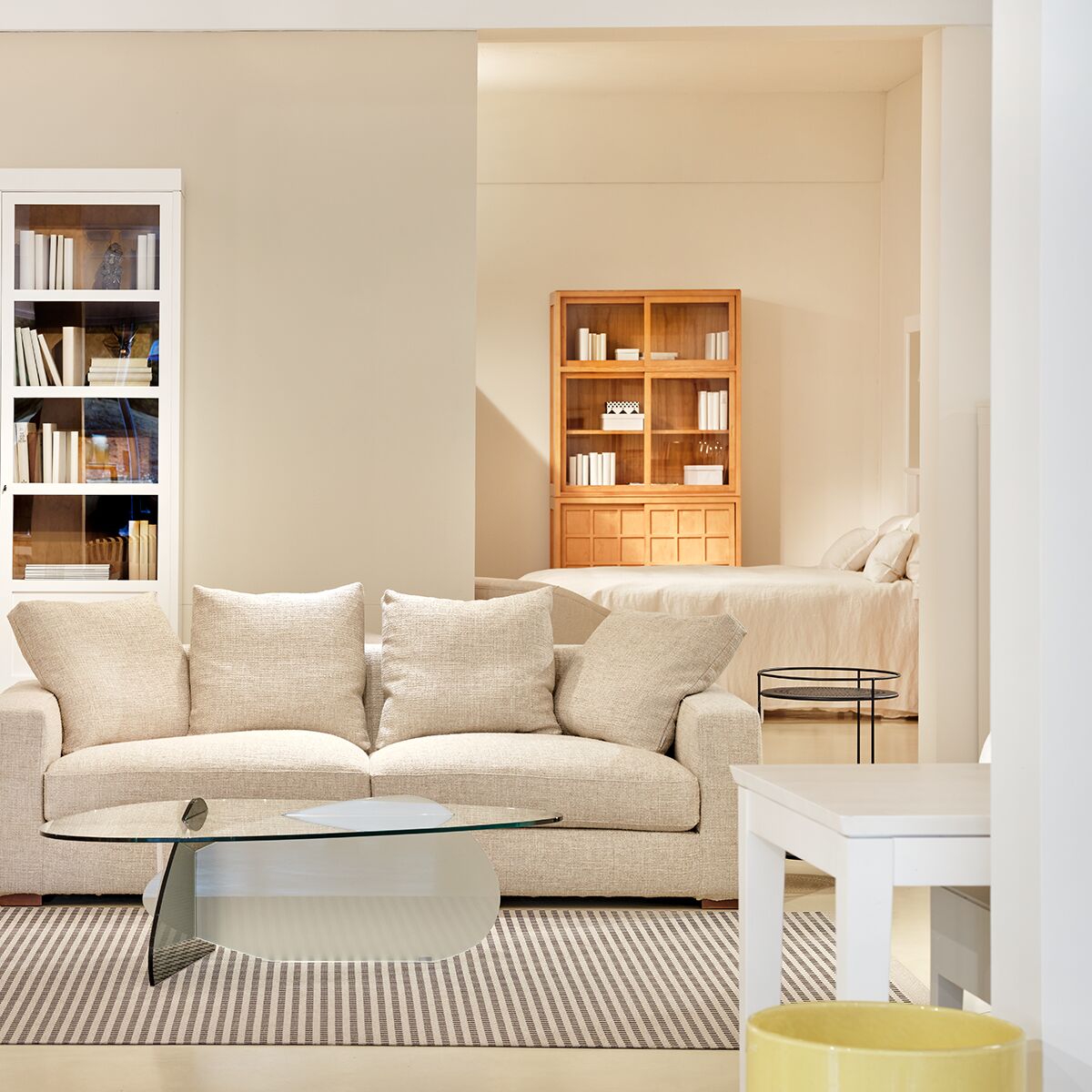 Modernes Wohnzimmer mit cremefarbenem Sofa, Glas-Couchtisch und Vitrine aus honigfarben lasiertem Massivholz mit klaren Glastüren. Im Hintergrund Schlafbereich mit weiterem Möbelstück aus passendem Holz. Helle, harmonische Atmosphäre und klare Linien.