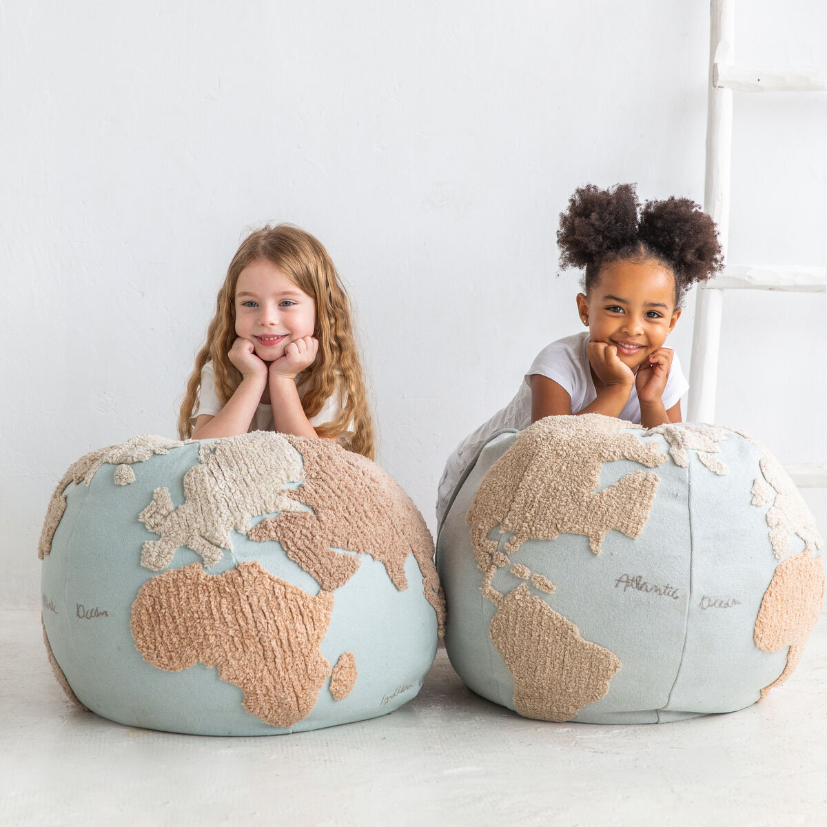 Zwei Kinder sitzen auf runden Sitzkissen mit Weltkartenmuster. Die Kissen sind in Beige und Blau gehalten. Die Kinder scheinen glücklich und bequem zu sein.