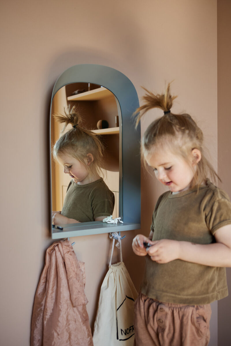 Ein kleines Mädchen steht vor einem blauen Wandspiegel in einem beige Kinderzimmer. Neben dem Spiegel hängen verschiedene kleine Gegenstände. Der Spiegel hat ein Regal und Haken darunter.