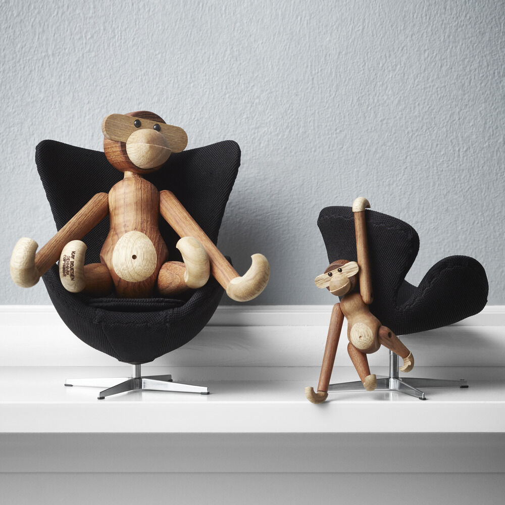 Zwei Kay Bojesen Holzfiguren „Affe“, gefertigt aus Teak und Limba, sitzen auf kleinen schwarzen Designerstühlen vor einer grauen Wand auf einem weißen Regal. Die Affen sind liebevoll gestaltet, verspielt arrangiert und wirken wie beliebte dänische Designikonen.
