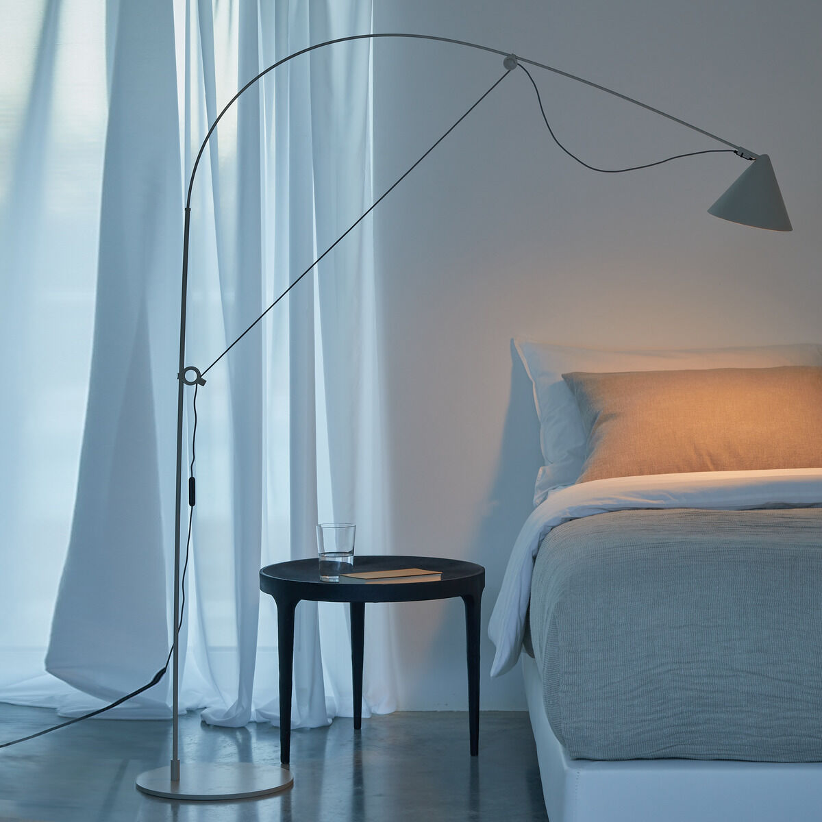 Eine moderne Stehlampe des Modells »AYNO« Silk neben einem Bett in einem Wohnzimmer mit minimalistischer Einrichtung. Die Lampe hat einen geschwungenen Schwenkarm und leuchtet warmes Licht auf das Bett. Daneben steht ein kleiner Tisch mit einem Glas und einer Wasserflasche.