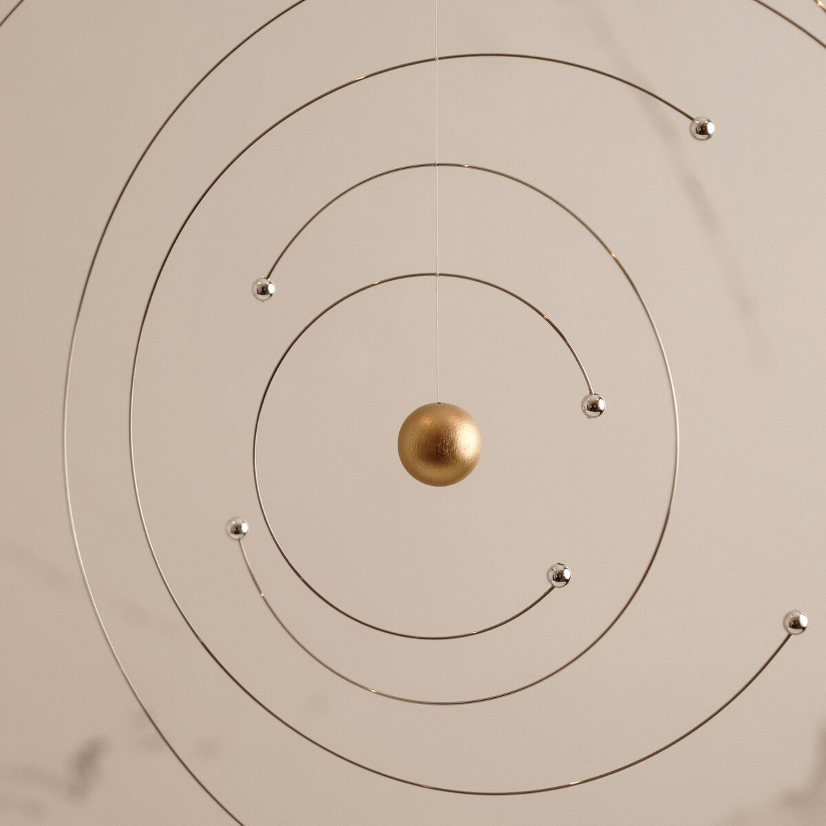 Ein künstlerisches Mobilé, das das Atommodell von Niels Bohr darstellt, mit einer goldene Kugel im Zentrum und mehreren Silberkugeln, die an feinen Drahtbahnen aufgehängt sind, auf einem beigen Hintergrund.