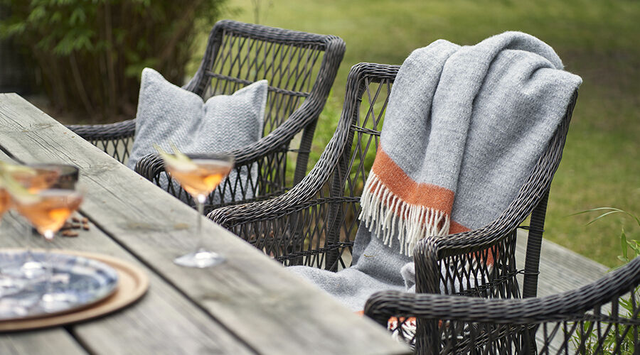 Grau-orangefarbene Decke mit Fransen auf schwarzem Gartenstuhl aus Rattan neben Holztisch mit zwei Gläsern Aperitif