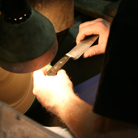 Eine Person schärft ein Käsemesser mit einem Holzgriff aus Olivenholz unter einer Lampe. Die Szene zeigt den Schärfvorgang mit einer rotierenden Schleifscheibe.