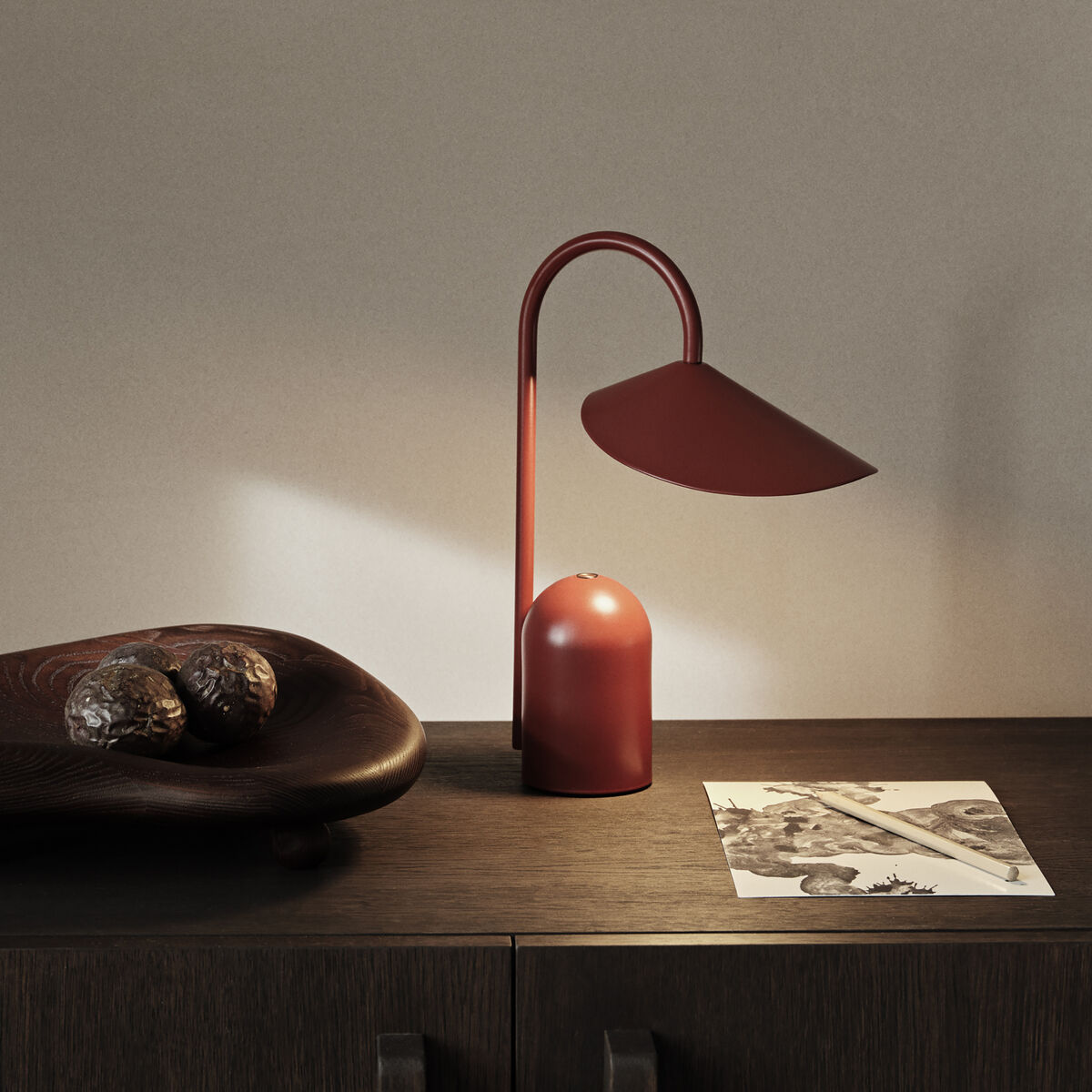 Ein Bild zeigt eine tragbare Lampe namens »Arum« auf einem dunklen Holzschrank. Die Lampe hat eine rote Farbe und ein modernes, minimalistisches Design. Neben der Lampe liegt ein Holztablett mit Dekorationsobjekten und ein Papier mit einer Skizze.