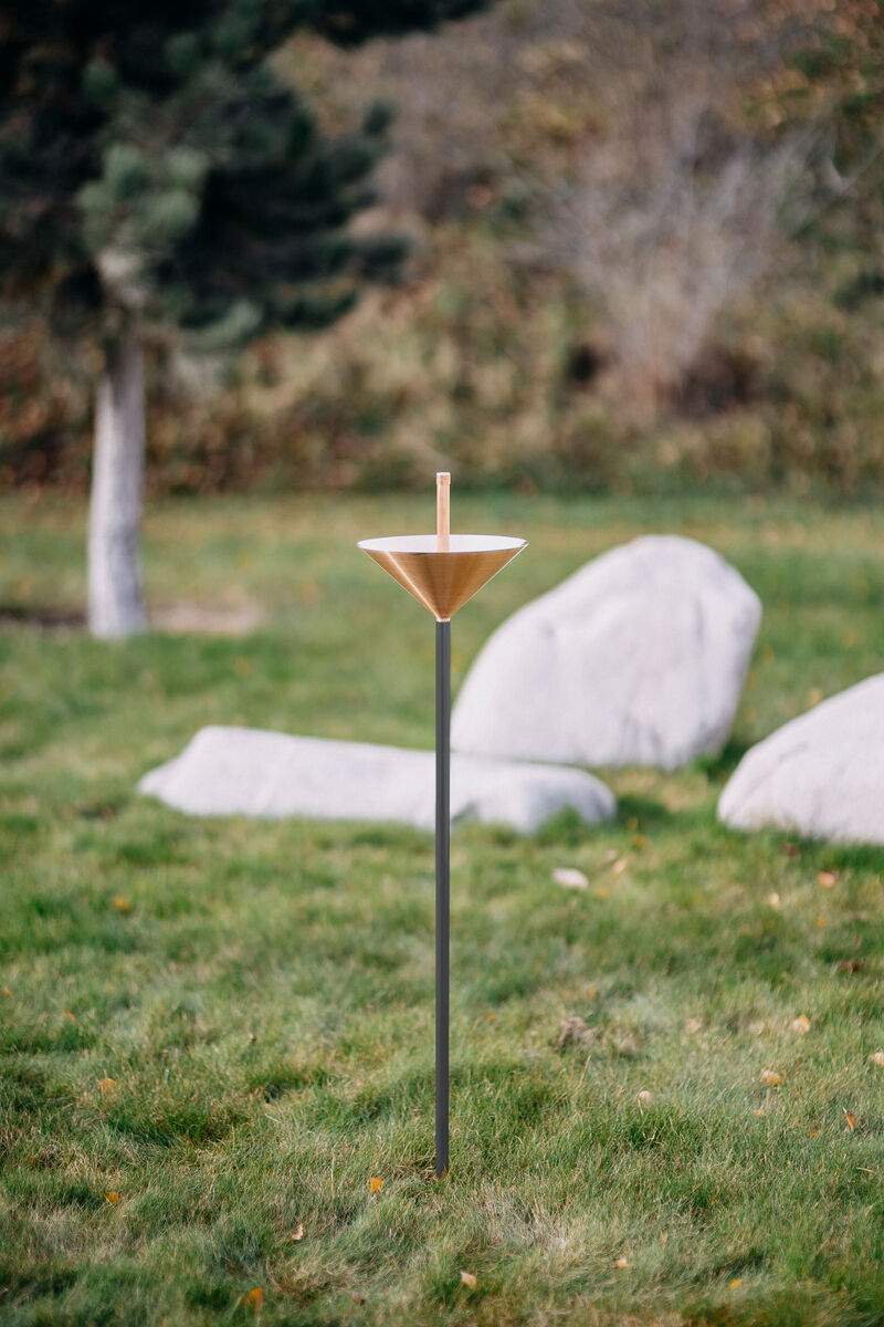 Regenmesser aus Kupfer mit langem Stab in einem grünen Garten. Im Hintergrund stehen Steine und ein Baum.