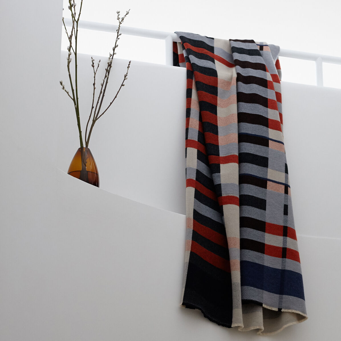 Eine Bauhaus-Stölzl Decke L mit grafischen Mustern in Weiß, Schwarz, Rot, Blau und Beige hängt über einem weißen Geländer. Im Hintergrund befindet sich eine Vase mit Zweigen.