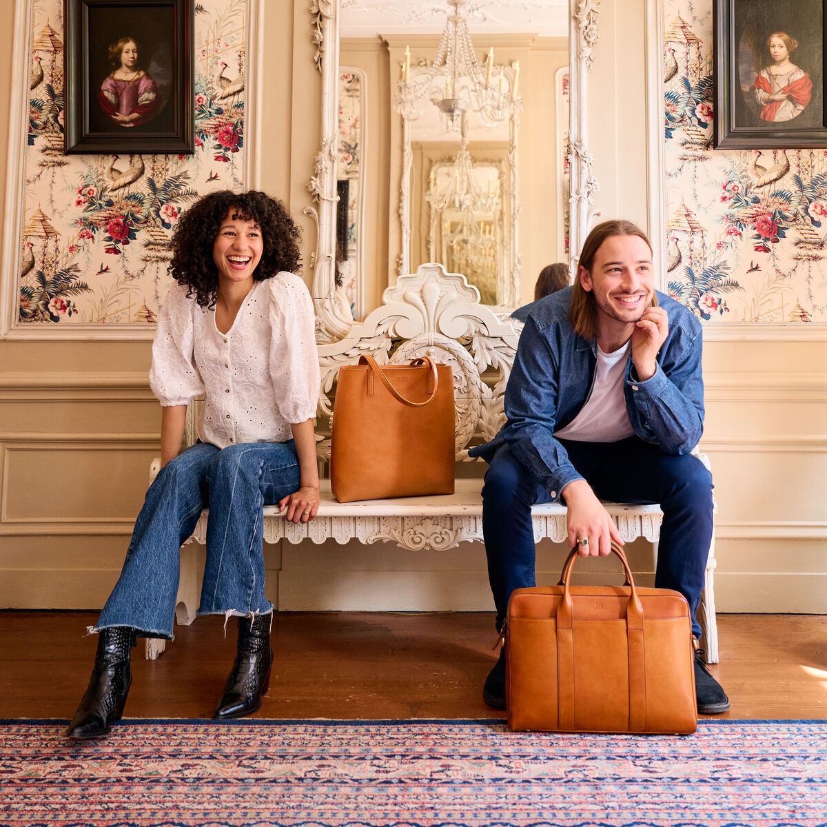 Zwei junge Erwachsene sitzen lächelnd nebeneinander auf einer stilvollen Bank in einem elegant eingerichteten Raum. Beide halten Ledertaschen des Modells »Harvey« in den Händen. Im Hintergrund sind eine große dekorative Spiegelwand und Gemälde zu sehen. Die Atmosphäre wirkt luxuriös und vintage.