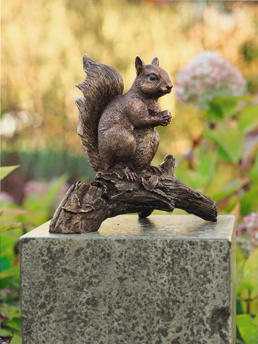 Gartenskulptur eines sitzenden Eichhörnchens mit einer Nuss in den Pfoten, auf einem Ast sitzend. Die Skulptur hat eine bronzefarbige Oberfläche und steht auf einem steinernen Sockel, umgeben von grüner Natur und unscharfem Pflanzenhintergrund.