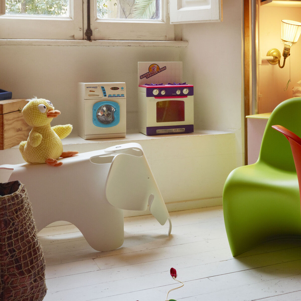 Ein farbenfrohes Kinderzimmer mit einem weißen Kinderhocker in Form eines Elefanten, bekannt als 'Eames Elephant'. Der Raum ist mit verschiedenen Spielzeugen und farbigen Möbeln dekoriert. Eine grüne und eine rote Stuhlfigur ergänzen das verspielte, moderne Design.