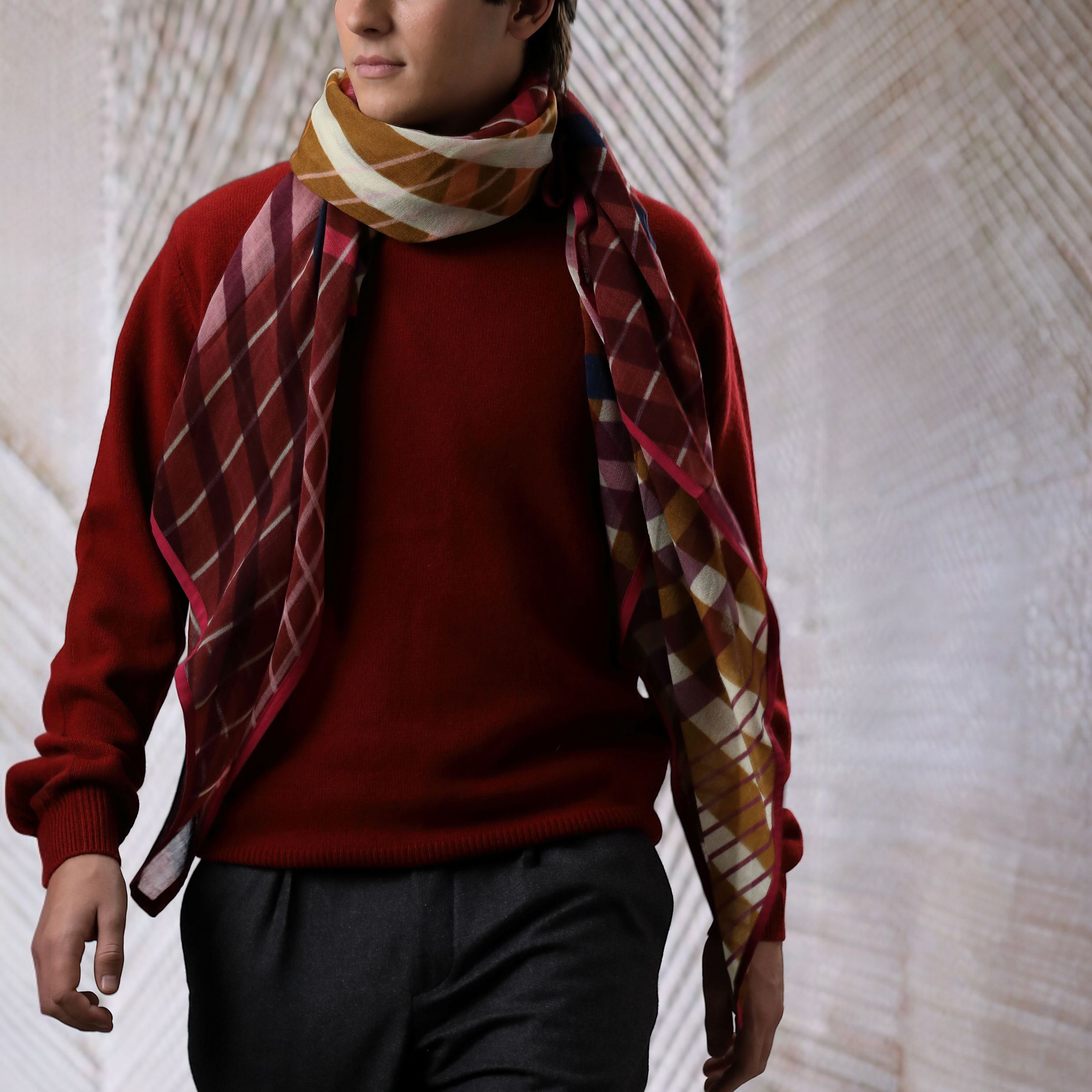Ein Wollschal im Modell ‚Indy‘ in Pink mit Karo- und Farbverlaufsmuster ist stilvoll um den Hals einer Person gebunden, die einen bordeauxroten Pullover trägt. Der Schal wirkt weich, wärmend und eignet sich besonders gut als modisches Accessoire für die kühleren Monate.