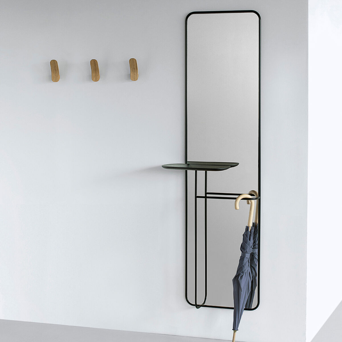 Ein moderner Spiegel namens 'Concierge' hängt an einer weißen Wand. Der Spiegel ist rechteckig und hat einen schwarzen Metallrahmen. Darunter befindet sich eine kleine Ablagefläche aus Glas und an der Seite hängt ein blauer Regenschirm. Weiterhin sind drei Holzhaken an der Wand montiert, die eine minimalistischen Stil unterstreichen.