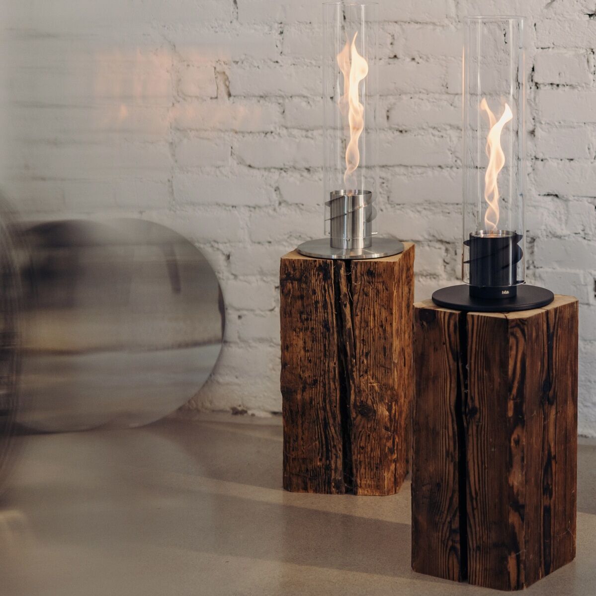 Zwei Tischfeuer mit hohen Glaszylindern auf Echtholzböcken, die vor einer weißen Backsteinwand platziert sind. Das Flammenbild sorgt für eine einladende und gemütliche Atmosphäre.