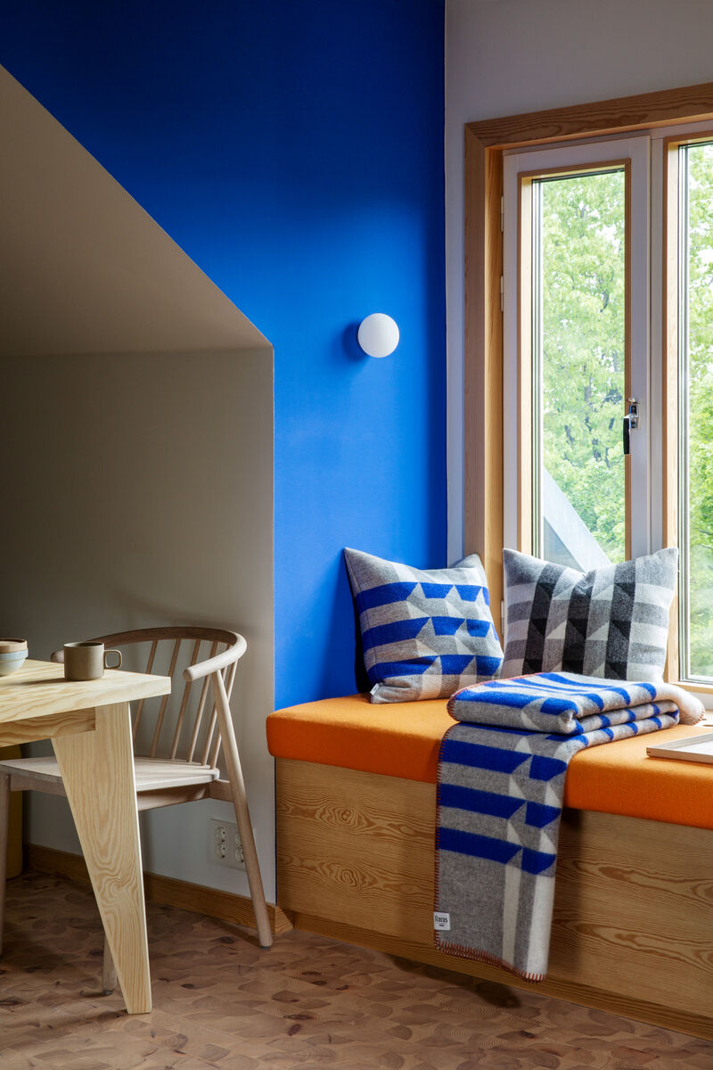 Ein gemütliches, modern eingerichtetes Zimmer mit einer Fensterbank dekoriert mit blauen und grauen Kissen sowie einer passenden Decke. Die Wand ist in einem auffälligen Blau gestrichen. Der Raum enthält auch einen Holztisch mit Teetassen und einen Holzstuhl. Draußen sind grüne Bäume zu sehen.