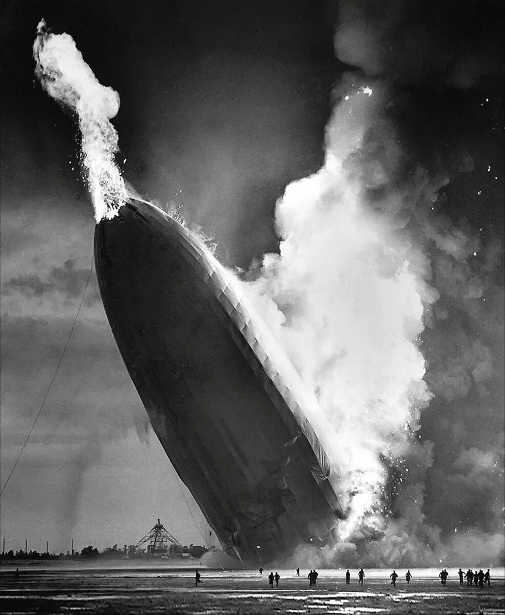 Ein schwarz-weiß Bild zeigt die brennende Hindenburg, ein großes Zeppelin, das zur Seite kippt, während Flammen und Rauch aufsteigen. Menschen stehen im Vordergrund und beobachten das Unglück.