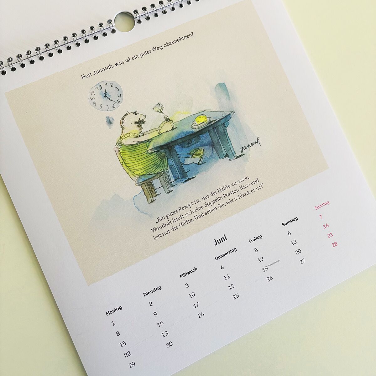 Der Wondrak-Kalender 2026 zeigt eine humorvolle Illustration mit einer Person an einem Tisch, auf dem ein Getränk steht, sowie einen Wandkalenderblatt für den Monat Juni. Die Seite enthält Wochen- und Tagesaufteilungen sowie einen Spruch oberhalb der Zeichnung. Die Farben sind überwiegend zart und pastellig gehalten.