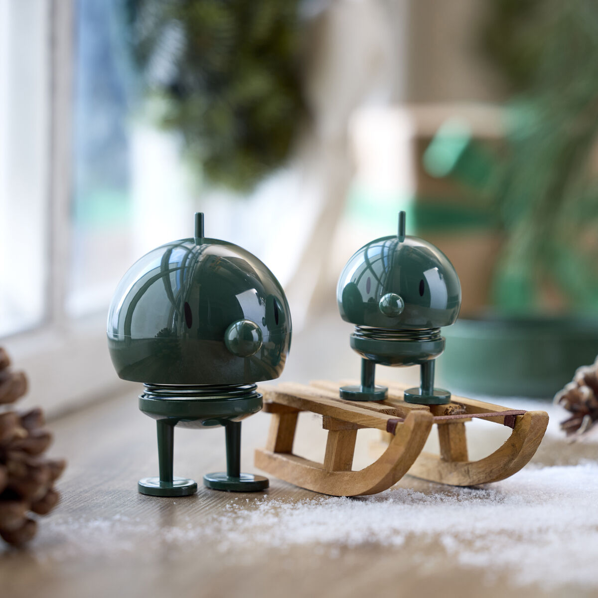 Zwei Hoptimist Bumble Figuren in Piniengrün aus ABS und Stahl stehen auf einem Holztisch, umgeben von Kunstschnee, Tannenzapfen und einem Holzschlitten. Im Hintergrund verschwommene Winterdeko und grüne Pflanzen, stimmungsvolles Licht.