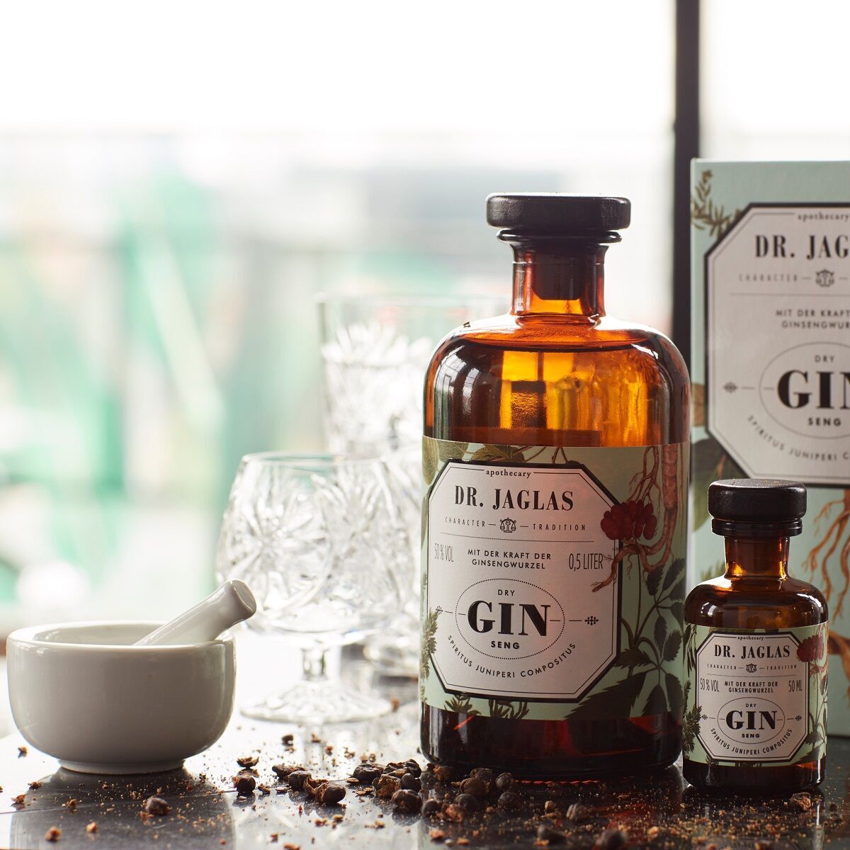 Die Abbildung zeigt eine Flasche »Dry GIN-seng Gin« von Dr. Jaglas in einem stilvollen Arrangement mit einer kleineren Flasche Gin, Gläsern und einem Mörser auf einem Tisch. Die Flasche hat ein elegantes Etikett mit grünen und weißen Elementen.