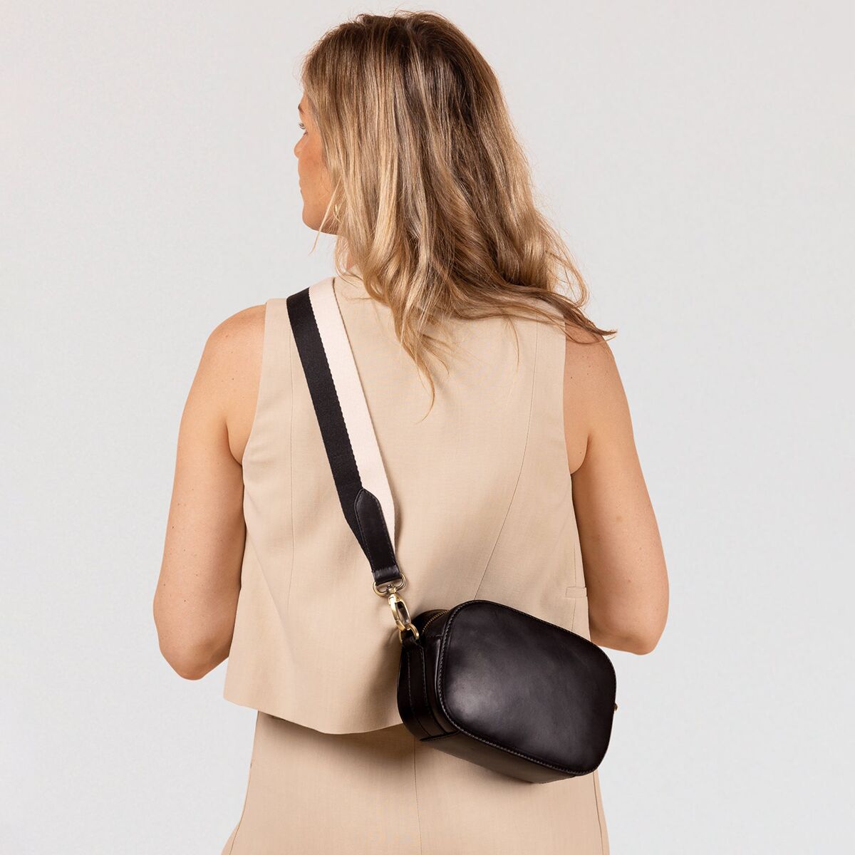 Eine Frau trägt eine mittelgroße, schwarze Umhängetasche aus Kunstleder mit beigefarbenem, breitem Schulterriemen über der Schulter. Die Tasche hat ein schlichtes, modernes Design und passt zu einem eleganten, beigen Outfit. Das Produkt bildet den klaren Fokus des Bildes, der Hintergrund ist neutral.