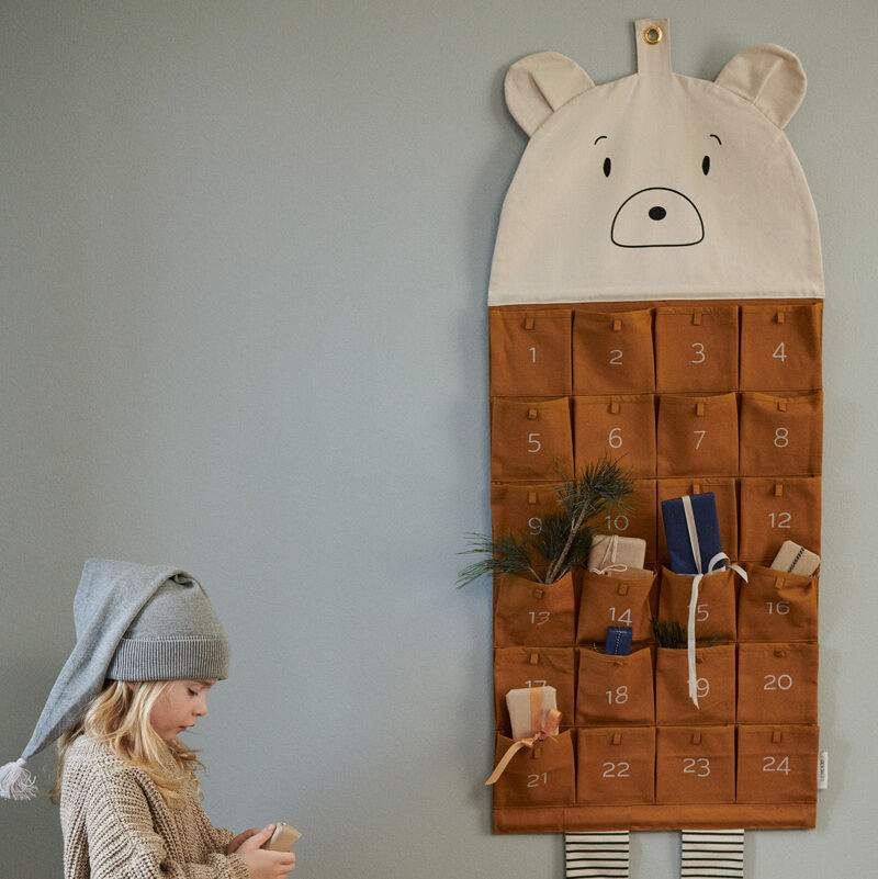 Ein Adventskalender in Form eines Tieres mit Bärengesicht hängt an der Wand. Der Kalender besitzt braune und nummerierte Fächer. Ein Kind mit grauer Zipfelmütze sitzt auf dem Bett und hält ein kleines Geschenk in der Hand. Das Bett ist mit buntem Bettzeug bedeckt.