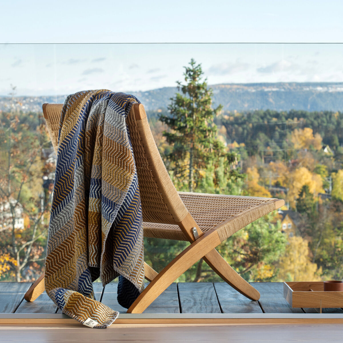Eine gemütliche Lammwolldecke in Blautönen und Gelbtönen hängt über einem Holzstuhl auf einem Balkon mit Blick auf eine bewaldete Landschaft.