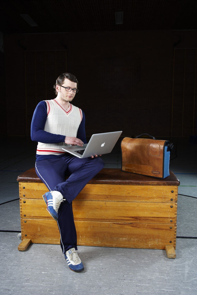 Ein Mann in Sportkleidung sitzt auf einem Holzturnkasten und arbeitet an einem Laptop. Neben ihm steht eine braune Ledertasche. Der Hintergrund der Szene ist eine dunkle Sporthalle.
