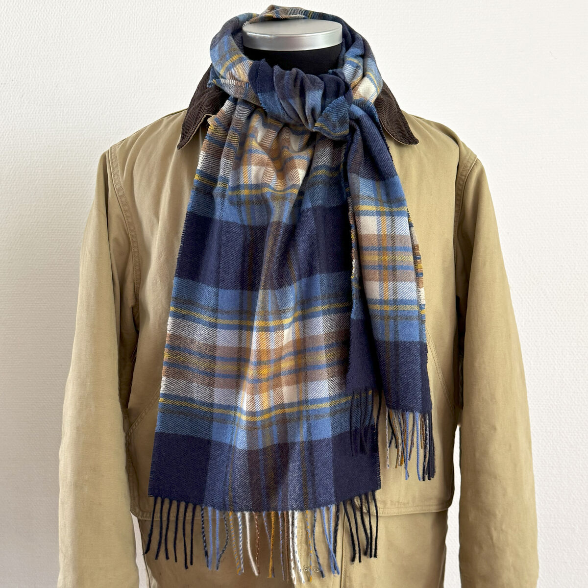 Ein beigefarbener Mantel auf einer Schaufensterpuppe, kombiniert mit einem karomusterten Kaschmirschal in verschiedenen Blautönen, Akzenten von Beige, Gelb und Weiß. Der Schal ist stilvoll um den Hals gebunden und sorgt für ein elegantes und warmes Erscheinungsbild.