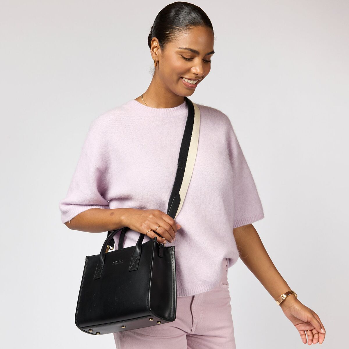 Eine Frau trägt eine schwarze Umhängetasche »Jackie Midi« an einer Schulter. Sie ist elegant gekleidet mit einem kurzärmeligen helllilafarbenen Pullover und einer passenden Hose. Die Tasche wirkt modern und geräumig, mit silberfarbenen Details und einem breiten cremefarbenen Schulterriemen. Der Hintergrund ist neutral, um den Fokus auf die Tasche und das Outfit zu lenken.