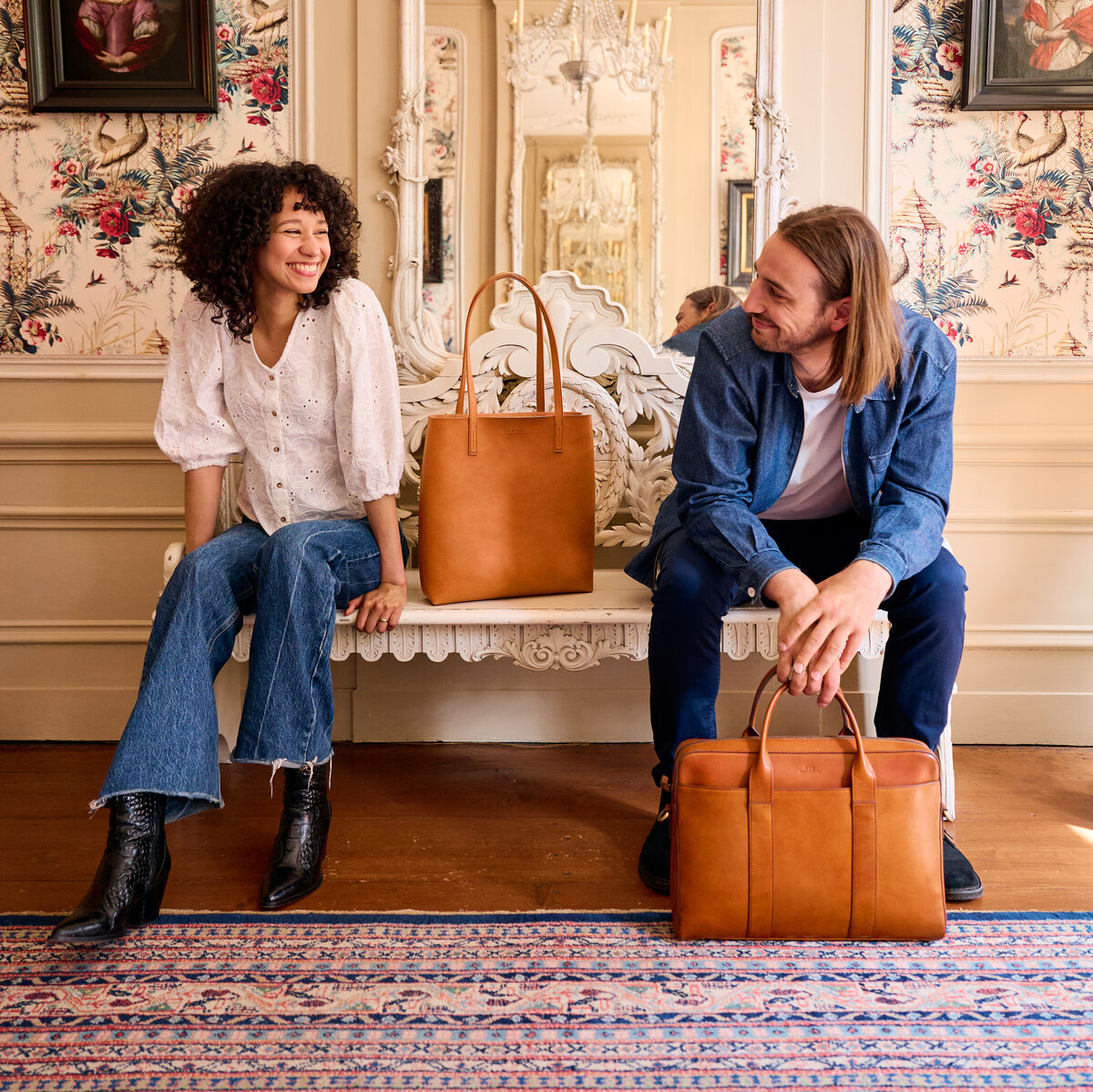 Zwei Menschen sitzen auf einer antiken Bank vor einem großen Spiegel in einem stilvoll dekorierten Raum. Beide halten stilvolle Kupferbraune Shopper-Taschen »Georgia«. Die Frau trägt eine weiße Bluse und blaue Jeans, während der Mann ein blaues Hemd und Jeans trägt. Der Raum ist mit floralen Tapeten und antiken Porträts dekoriert.