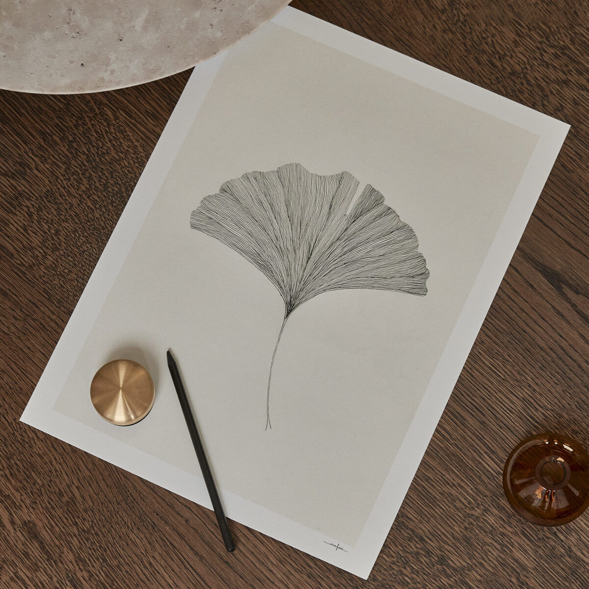 Ein Kunstwerk von Ana Frois mit einem Ginkgo-Blatt. Das Bild ist gerahmt und zeigt die feine Struktur des Blatts. Der Hintergrund ist braun und minimalistisch dekoriert, mit einem goldenen runden Objekt und einem dunklen Stift an der Seite.