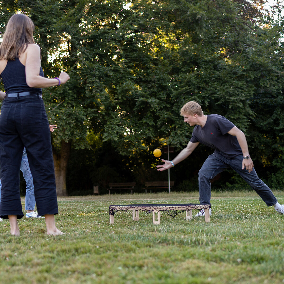 Personen spielen auf einer Wiese ein Freizeitspiel mit einem Holzrundnetz und einem gelben Ball. Im Hintergrund sind Bäume zu sehen. Die Szene wirkt sommerlich und entspannt.